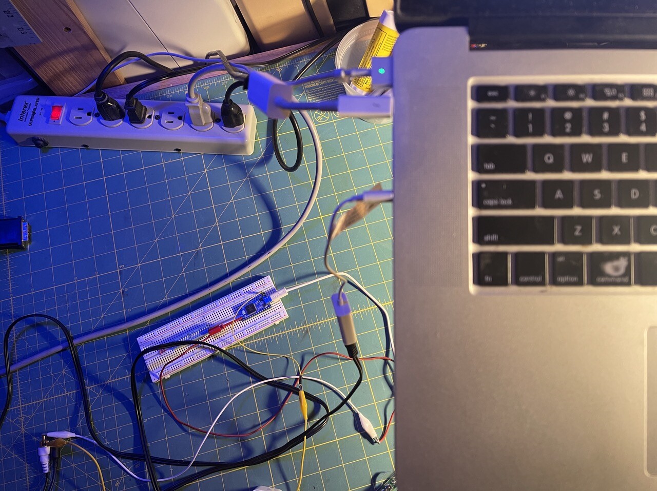 In the foreground on the right is half the keyboard of a 2009 MacBook. Below on the left is a protoboard with a teensy, wires, and a power strip all on a green workbench mat. 