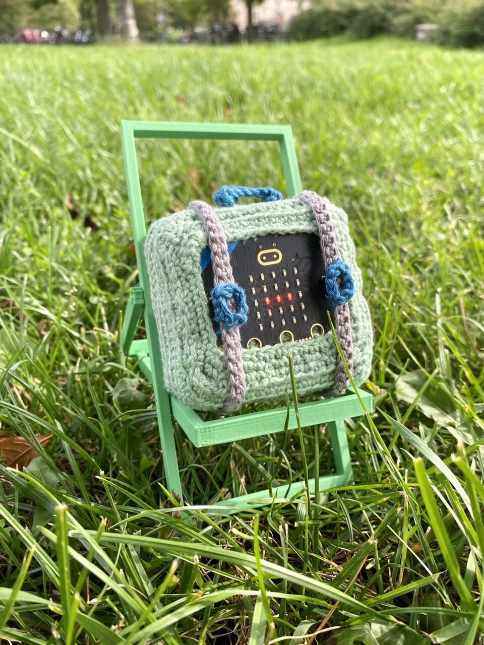 A pastel green crocheted travel suitcase with grey straps and blue handles. The front of the case is embedded with a Micro:Bit, which is an electronic microcontroller board with a grid of LEDs. Some of the LEDs are glowing and the straps cover the buttons __on the board. The suitcase rests on a 3D printed deck chair laid on some grass in a park.