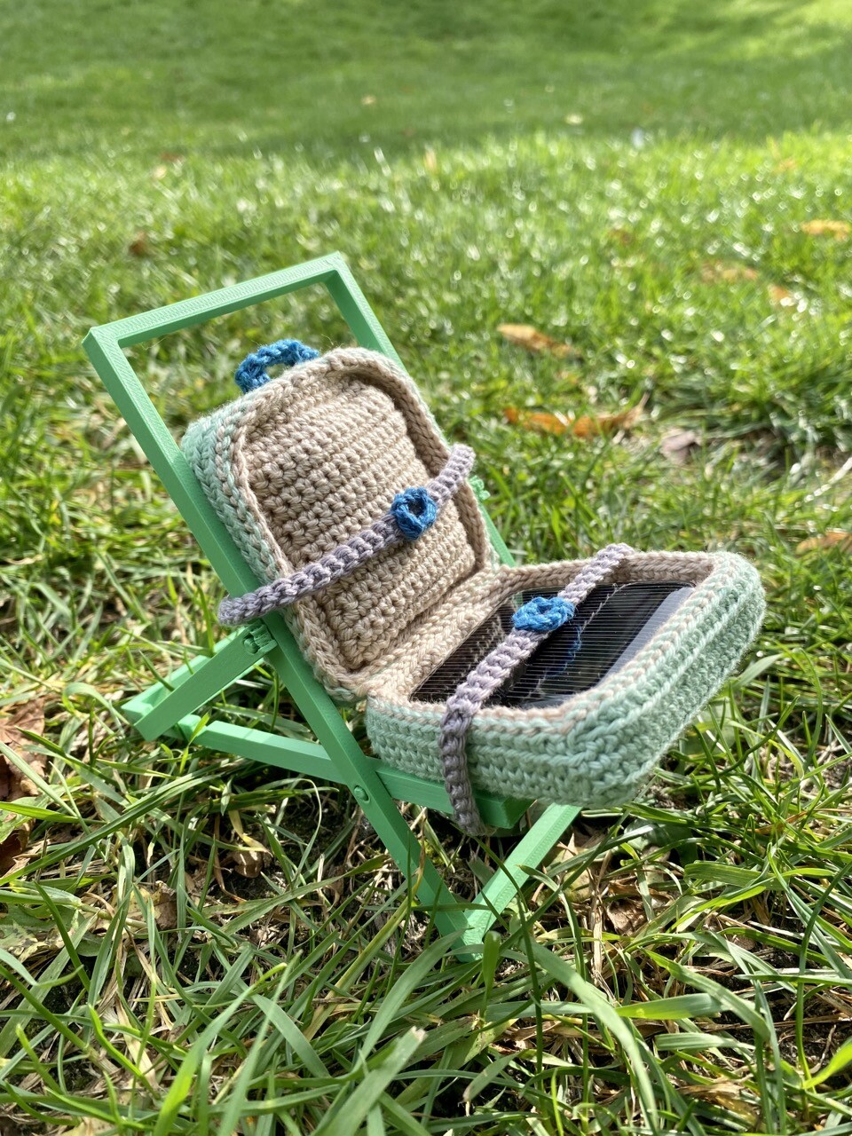 A pastel green crocheted travel suitcase with grey straps holding the suitcase in place on a 3D printed deck chair. The suitcase is opened, revealing a solar panel inside. The panel is left to charge under the sun. The background is grass in a park.