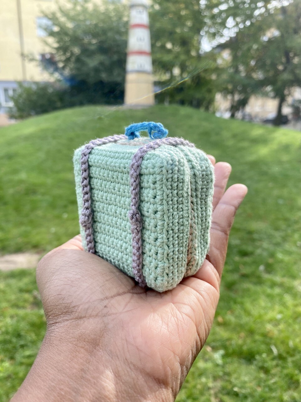A pastel green mini travel suitcase in crochet. My hand is holding the case and showing it’s backside. The case has gray straps and a blue handle.