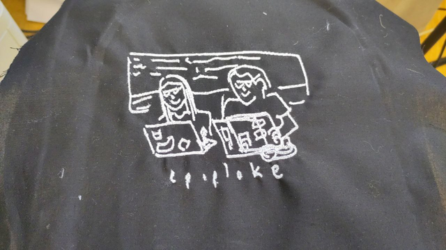 Machine-embroidered line drawing of two smiley people on laptops with 'epiploke' underneath. White thread on black cotton.