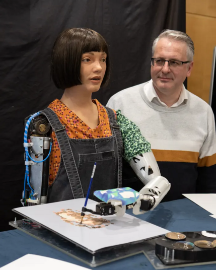 Older man looking a bit creepily at his young female-presenting 'ai' robot painter.